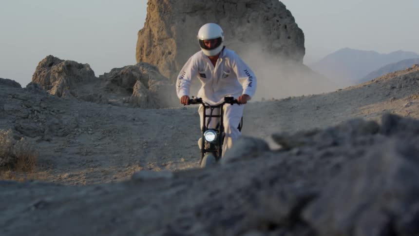 A cyclist dressed in white and wearing a white helmet rides a motorbike in a desert with large rock formations
