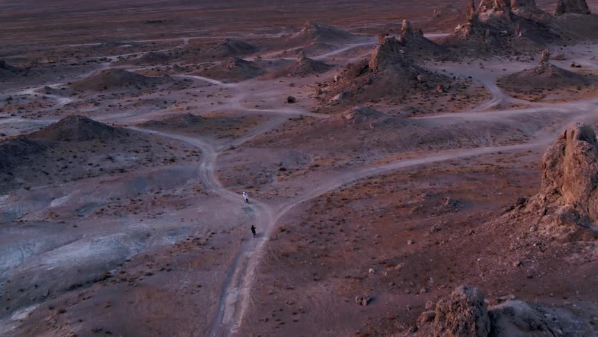 Two people riding motorcycles on a dirt road in a desert landscape with the sky and clouds in the background