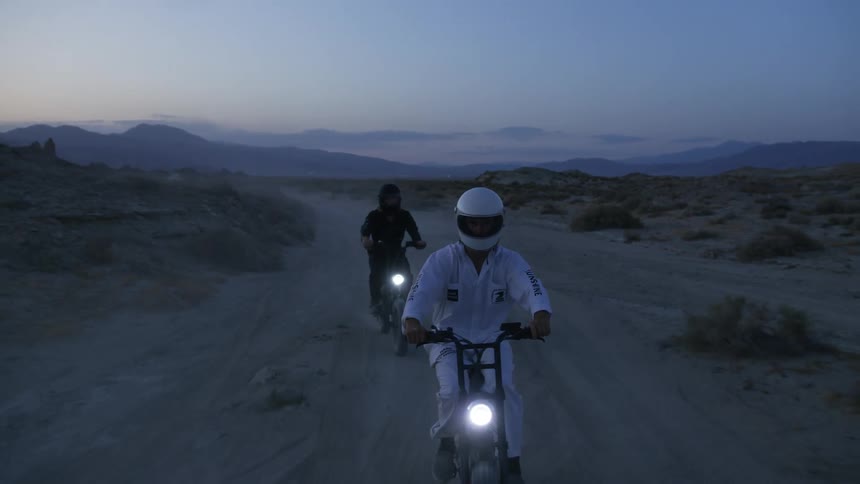 Two people are riding electronic bicycles on a sandy path in the desert wilderness at dusk