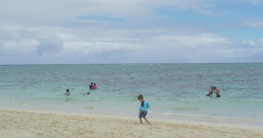 A boy walks in the sand as others are in the water