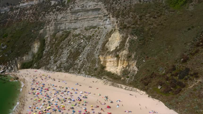 People mill about beach as others move about parking lot on top of cliff under deep blue sky