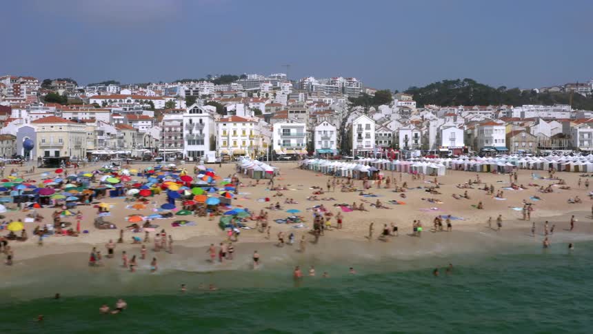 A beach with people umbrellas water and waves with buildings in the background