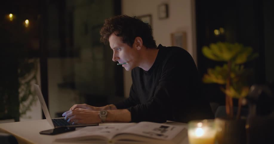 A man is sitting at table in a home while working on a laptop computer at night with candles burning