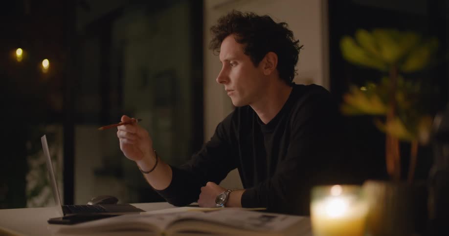 A man is stilling at a table with a computer in front of him as he plays with his pencil