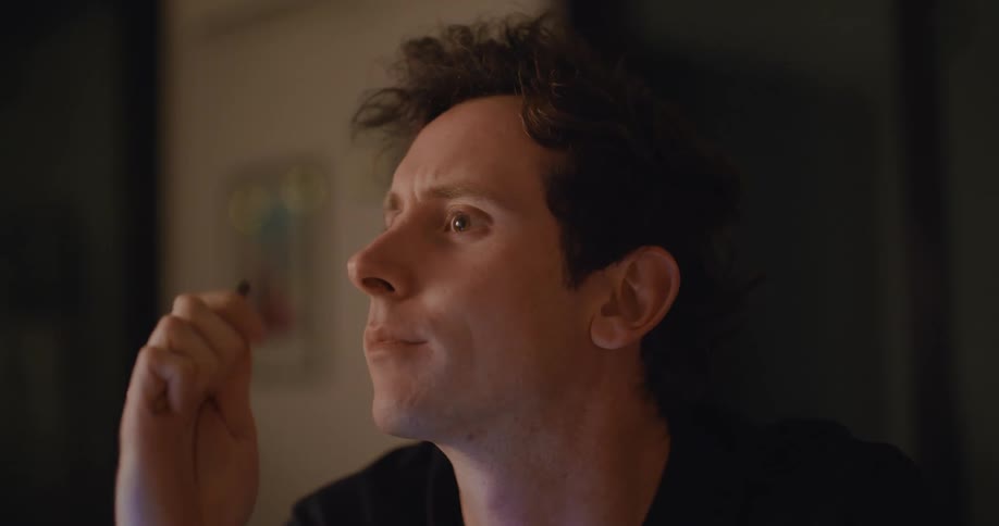 A man is sitting at a table with a laptop while at home thinking intensely about something at night