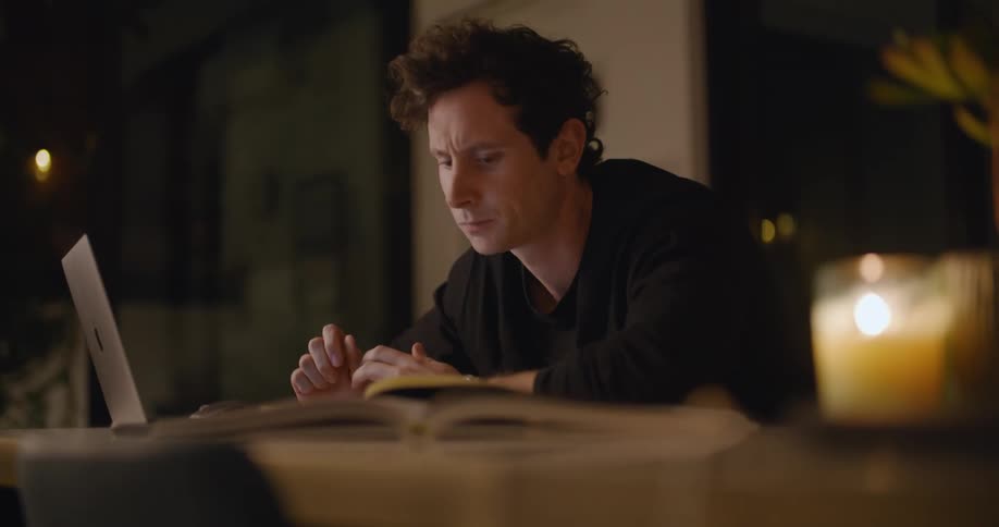A man sitting at a table reads the screen of a laptop before writing in a notebook