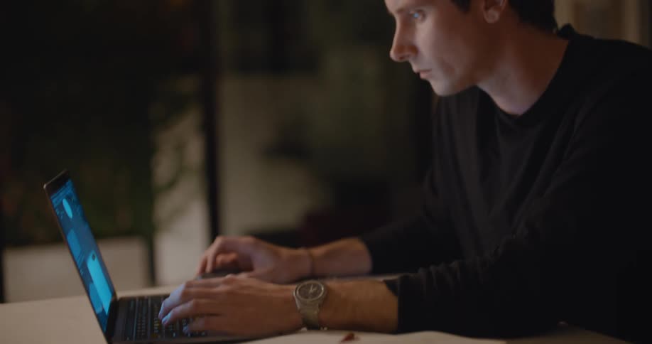 A man sitting at a table writes in a notebook before turning to use his laptop