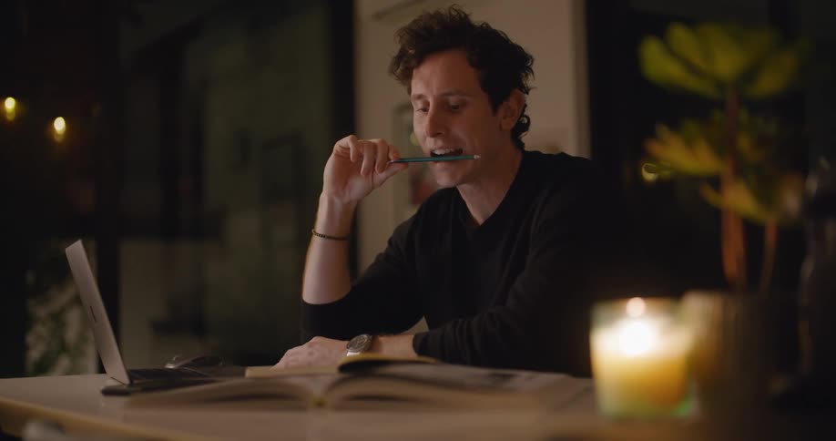 A man sitting at a desk with a laptop and books fidgets with his pencil and rubs his face in tired frustration