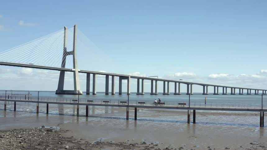 Cyclists pedal across a small bridge along a beach next to a large bridge carrying traffic