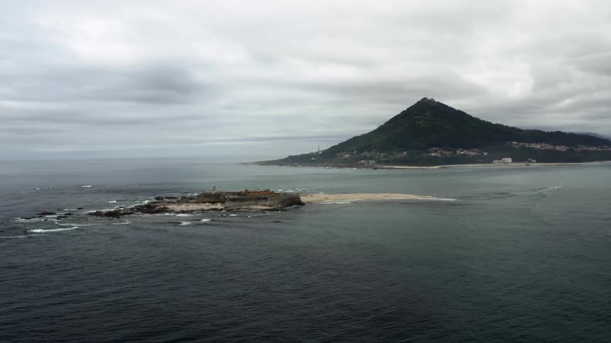 Ocean waves splash against the rocks and sand of a small island off the coast of hilly land