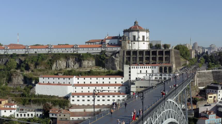 Camera zooms by a crowd walking on a bridge onto a few buildings situated on a cliff in a city around midday
