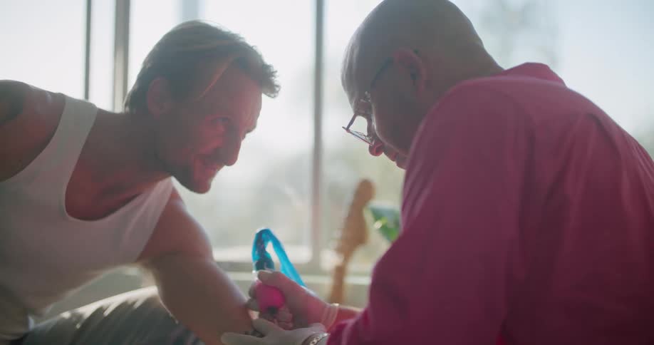 A man wearing a bright pink shirt is giving another man a tattoo