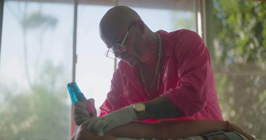 A man wearing a bright pink shirt is giving someone a tattoo on their back