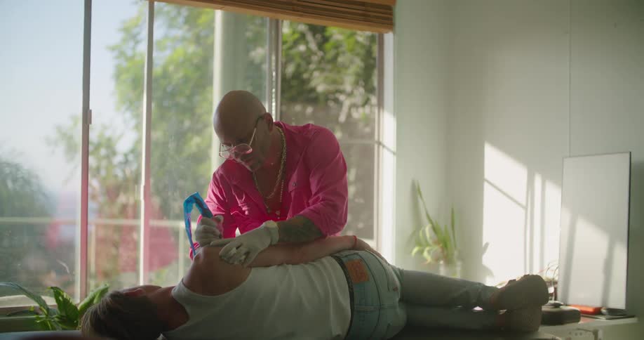 A man in a room with gloves giving a tattoo to another man with a wall and window in the background