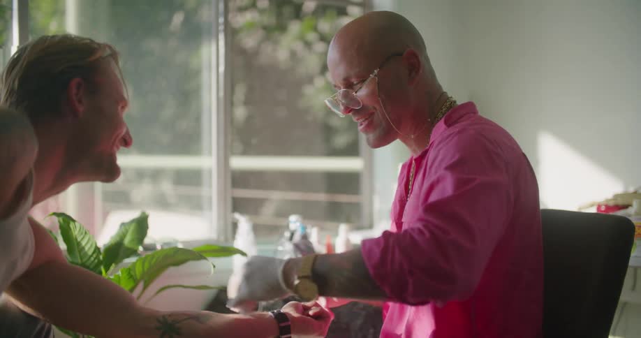 Tattoo artist with golden glasses necklace and wristwatch applies tattoo on mans upper wrist in tattoo shop