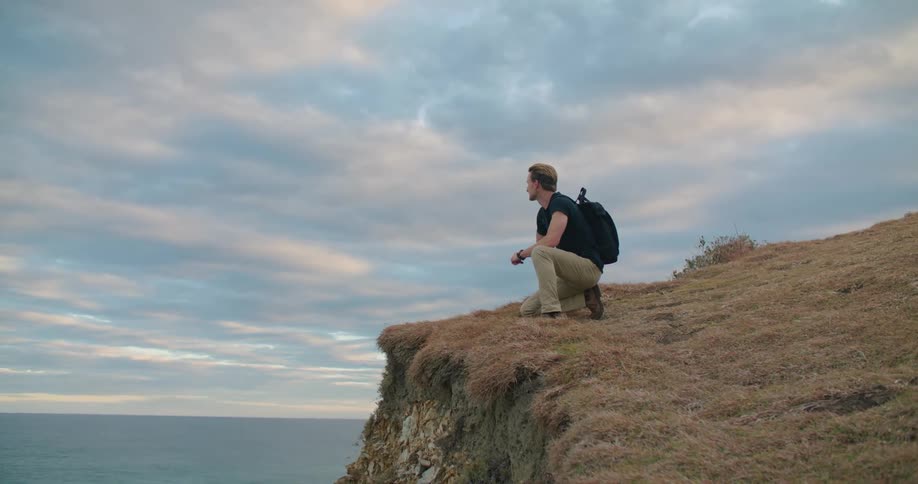 A man on one knee at the edge of a cliff overlooking the ocean below