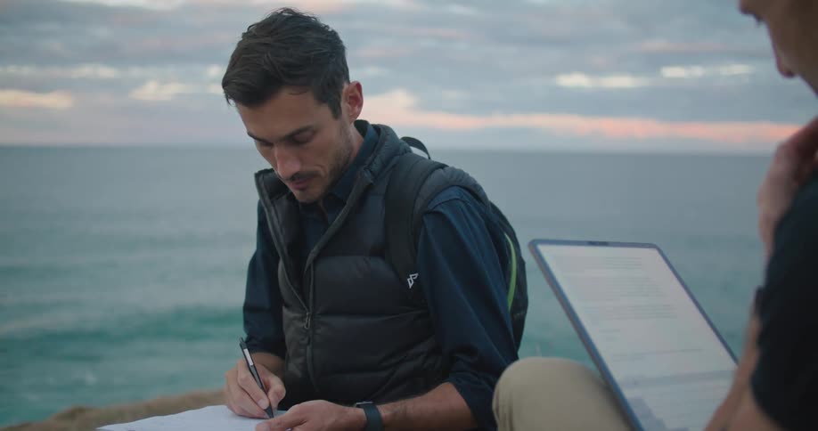A man sits on the shore and looks at a tablet as he talks with another man who takes notes
