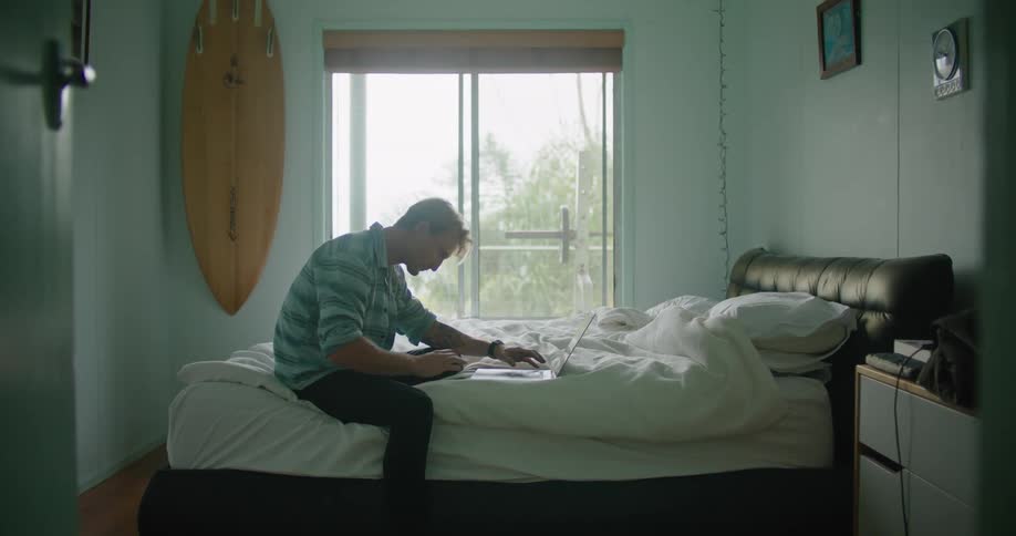 A man is sitting on a bed as he types on a tablet