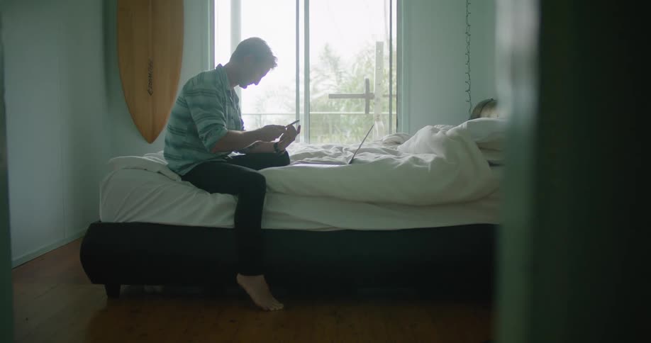 A man sitting on his bed holding his phone tapping with his laptop open in front of him