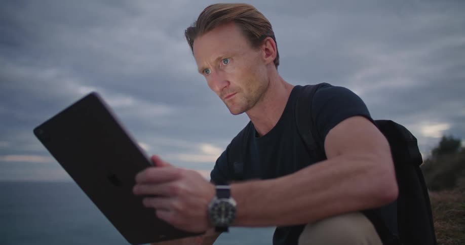 A man looks at his tablet as he sits next to the ocean