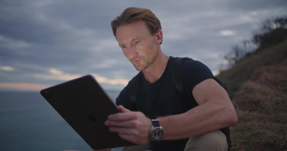 Man enters data using tablet while crouching with ocean and red tinted horizon in background