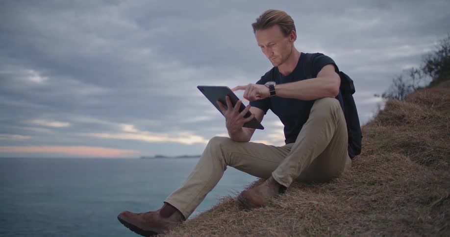 A man is sitting on a hill holding a tablet in one hand and navigating it with the other hand