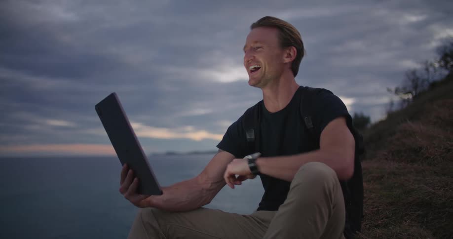 A man is sitting on a hill having a video conference on a computer tablet and laughing as he talks