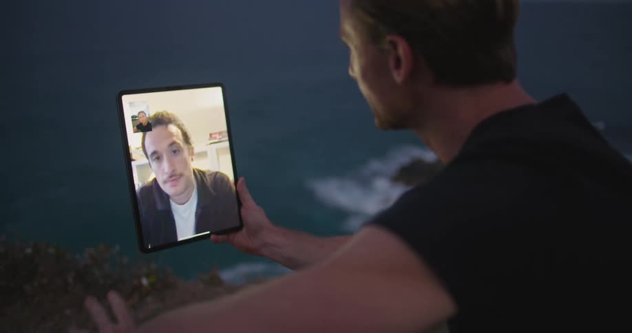 A man sitting on the shore holds a tablet and video chats with another man