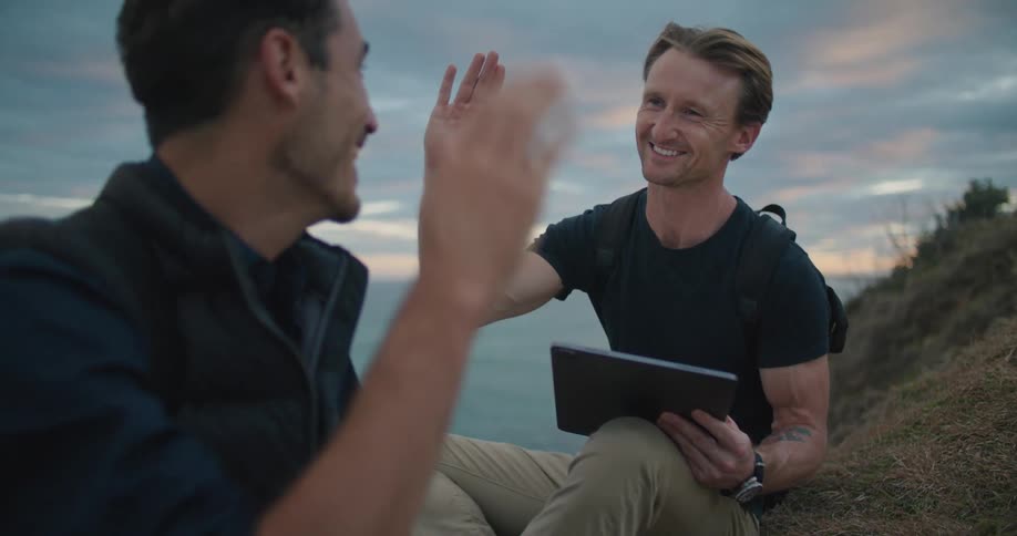Two men are sitting and talking on a grass covered hill along the shore and one of the men is holding a computer tablet