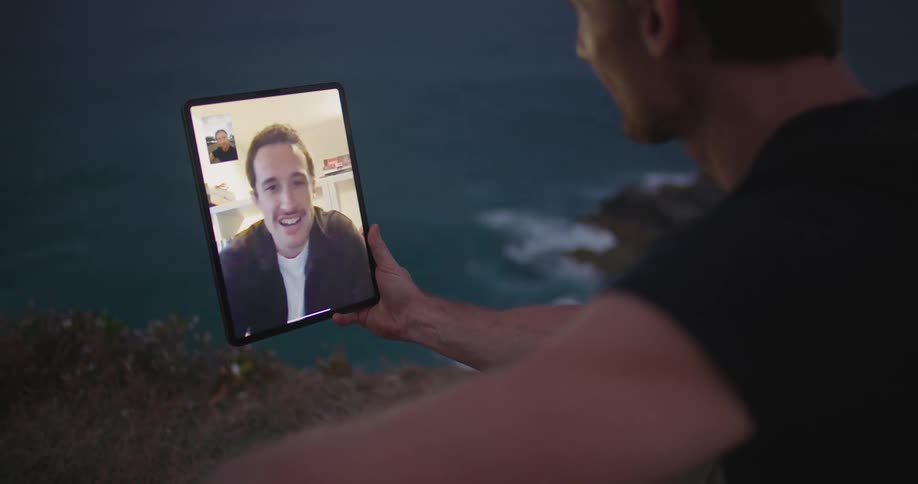 A man sits on a cliff overlooking a coastal area as he talks to another man on the tablet