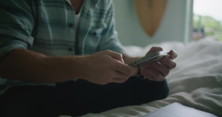 A man is sitting on a bed looking worried as he looks at a cell phone