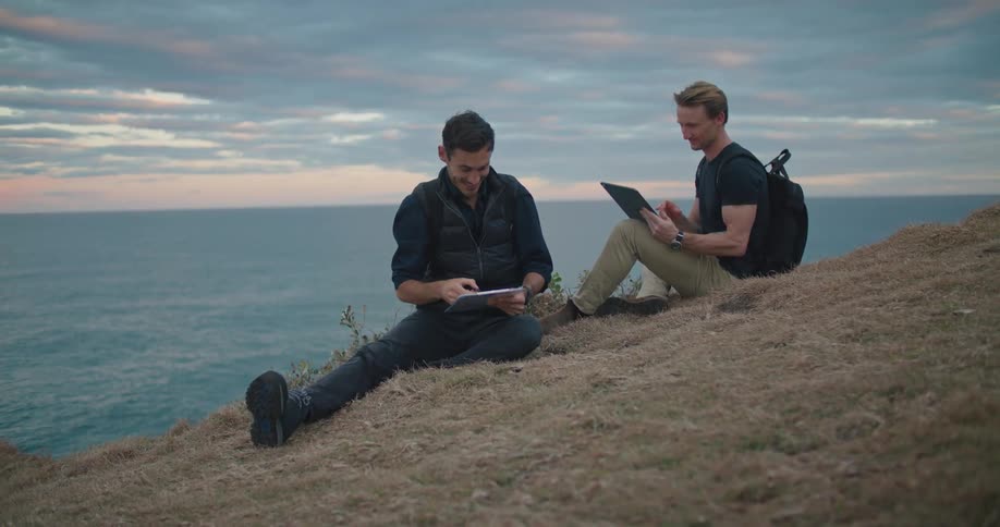 Two men are sitting and talking on a grass covered hill along the shore and one of the men is holding a computer tablet and the other is holding a clipboard