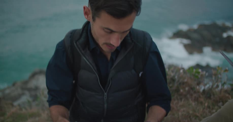 Two men are on a cliff overlooking the waterfront as they look at a tablet and papers on a clipboard