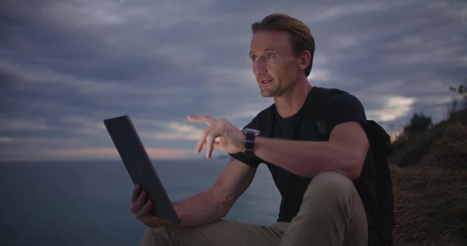 Man sits on shore near hill talking via tablet with red sky over ocean on horizon in background