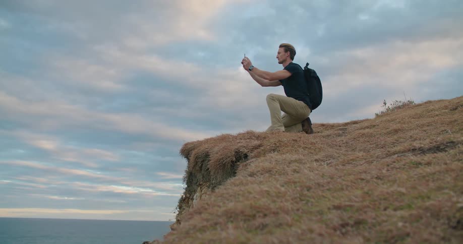 A man is kneeling at a cliff overlooking the ocean and taking pictures with a cell phone