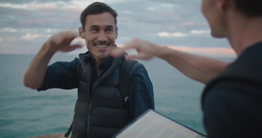 Two men sitting on a ridge overlooking the ocean working on a tablet and clipboard high five each other