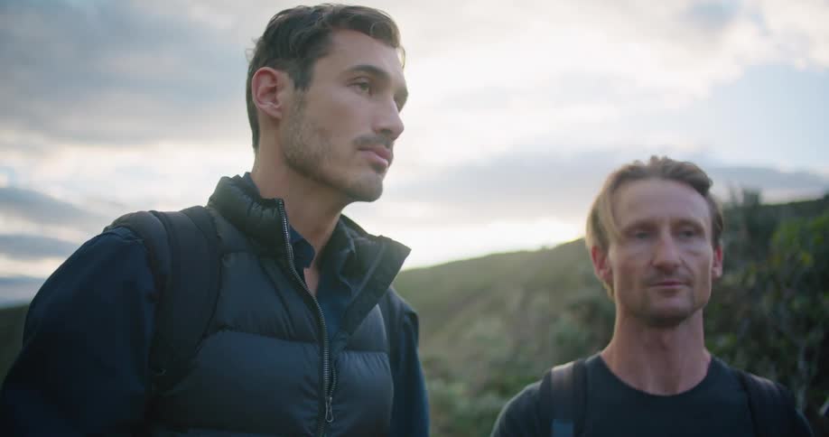 Two men with backpacks with a hill the sky and clouds in the background