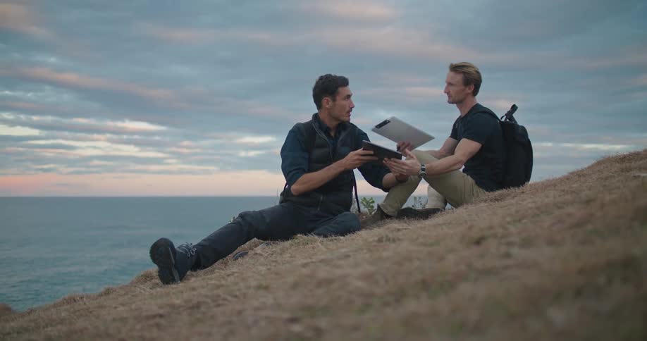 Two men sit on a hill on the shore and look at a tablet and talk and take notes