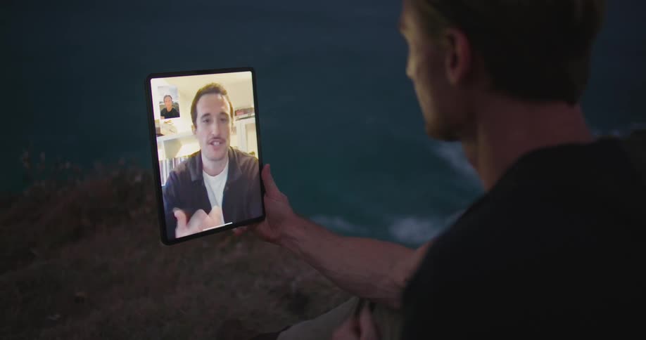 Blonde man with black t shirt makes video call on tablet sitting on the edge of cliff overlooking wavy sea