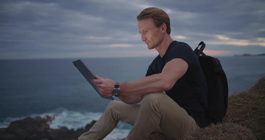 A man uses a computer tablet while sitting on a cliff above the ocean on a cloudy evening