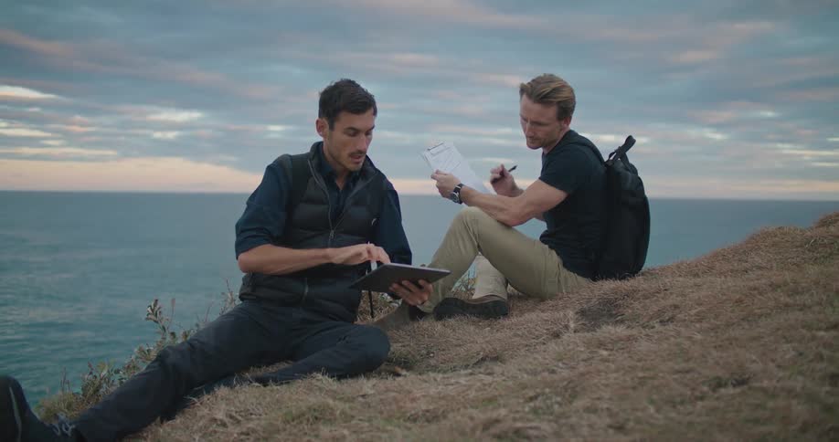 Blonde man with dark blue shirt backpack and brown pants sits on hillside with man backpack vest and shirt and jeans using tablet and pointing out on the hill overlooking sea