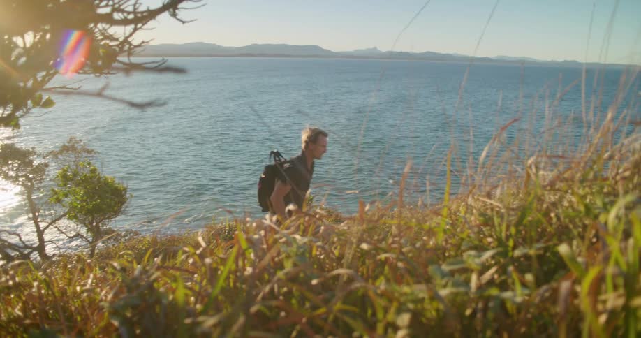 A man is looking through binoculars then hiking up a grassy hill overlooking the ocean