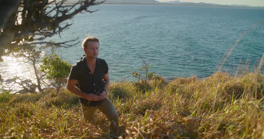 Blonde man with backpack dark blue shirt and brown pants walks uphill overlooking the sea as the sun shines brightly