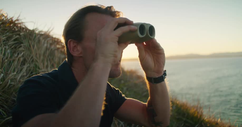 A man is sitting in the grass on a hill along the shore looking through binoculars as the sun shines brightly behind him