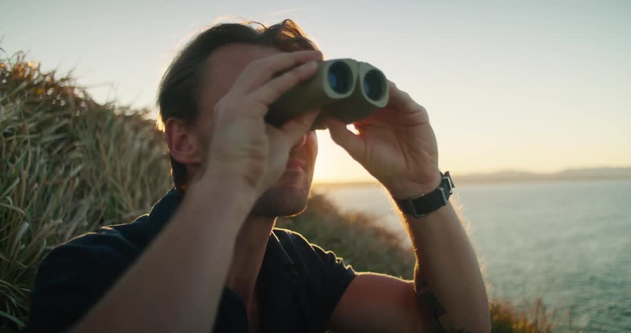 Man sits on shore near water observing with binoculars and making notes on tablet