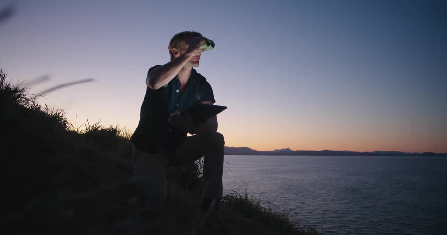 Man kneeling on a hill near the ocean uses binoculars and takes notes on a tablet at sunset