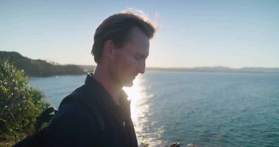 A man with a backpack by a lake looking through binoculars with the sky and clouds in the background