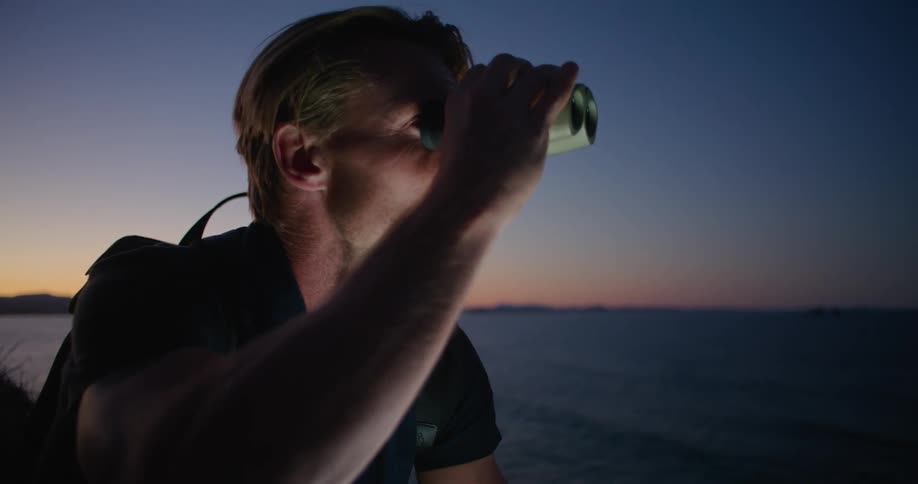 A man is sitting along the shore holding a computer tablet in one hand and binoculars in the other hand