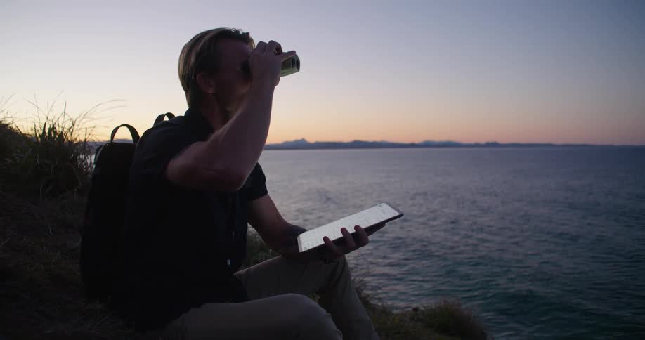 A man taps on his tablet and looks through binoculars while sitting on a ridge overlooking the ocean