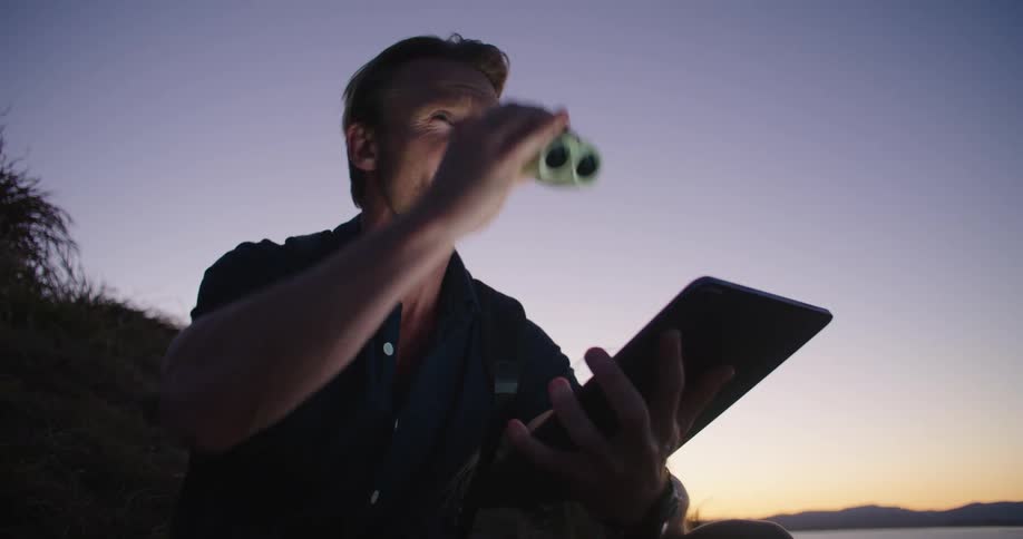 A man is sitting and using an electronic tablet and binoculars on a hill overlooking the ocean